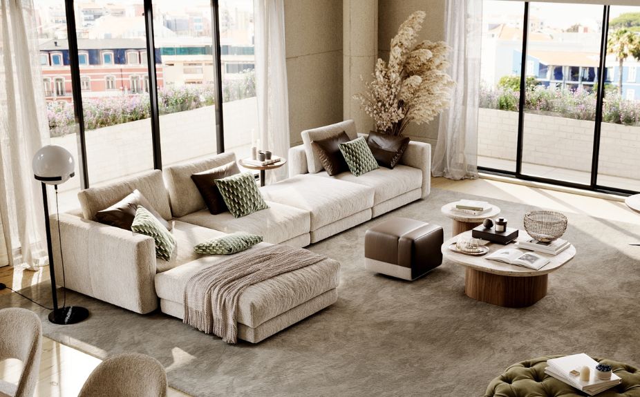 A large modular sofa in beige tones with green and brown cushions, a dark leather puff, and organic coffee tables.